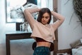 Modern charming young woman in a stylish pink sweatshirt in fashionable blue jeans is resting sitting on a chair in a cozy bedroom Royalty Free Stock Photo