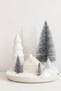 Modern christmas scene, miniature snowy village on white table. Merry Christmas! Stylish little Christmas trees and house. Winter