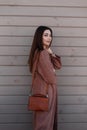 Modern comely young woman with beautiful hair in long youth spring coat with leather fashionable handbag stands near wooden