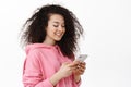 Modern curly girl texting, chatting on smartphone, smiling and looking at mobile phone, using application, standing Royalty Free Stock Photo