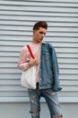 Modern cute young man in fashion pink sweatshirt in stylish denim jacket with vintage fabric shopper stands near white wall in Royalty Free Stock Photo