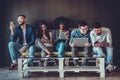 Diverse students using gadgets, sit on sofa in line on grey wall background indoors