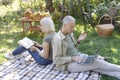 Modern elderly spouses resting in garden, man using laptop computer while his wife reading book, sitting back to back Royalty Free Stock Photo