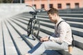 Modern employee sitting on stairs and networking outdoors with takeaway coffee and bike