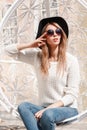 Modern european young hipster woman in a luxurious hat in stylish jeans in a knitted sweater in sunglasses posing sitting Royalty Free Stock Photo