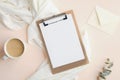 Modern feminine workspace with cup of coffee, blank clipboard, white blanket, envelope and eucalyptus leaf on beige background.