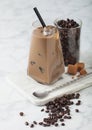 Modern glass of iced coffee with milk on marble board with jar of coffee beans and salted caramel and long spoon on light Royalty Free Stock Photo