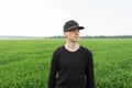 Modern handsome young man in a fashionable black cap in a stylish t-shirt is resting standing in a field among green grass. Royalty Free Stock Photo