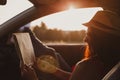 Modern hipster girl resting in a car and reading a map. Woman with feet on car door. Feet outside the window at sunset Royalty Free Stock Photo