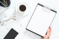 Modern home office desk workspace with blank paper clipboard in woman hand, coffee cup, glasses, smartphone, stationery on white Royalty Free Stock Photo