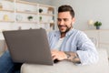 Modern job lifestyle. Happy tattooed man sitting on sofa at home and working on laptop