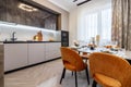 Modern kitchen combined with the dining room in dark and light colors, with orange chairs and glass cabinets for dishes