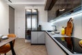 Modern kitchen combined with the dining room in dark and light colors, with orange chairs and glass cabinets for dishes