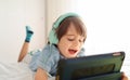 Modern Little boy in headphones is using a digital tablet and smiling while lying on his bed at home - Cute kid in a blue shirt