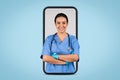 Modern medical services. Happy young female latin doctor in blue uniform posing with folded arms in big cellphone screen