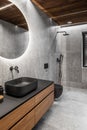 Modern minimalist bathroom interior with stone grey tiles, wooden furniture, black sink and toilet and round mirror. Royalty Free Stock Photo