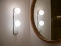 Modern mounted wall light lamp near the round shape wooden mirror on white wall background in the bathroom.