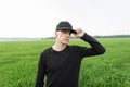 Modern nice young man in a fashionable black cap in a stylish black shirt posing among green grass in a field outside the city. Royalty Free Stock Photo