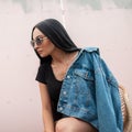 Modern pretty young woman with black hair in sunglasses in a stylish denim jacket in a t-shirt sitting on a chair near a pink wall Royalty Free Stock Photo