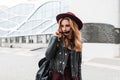 Modern pretty young woman hipster in a vintage purple hat in a fashionable jacket with a leather backpack with sunglasses stands Royalty Free Stock Photo