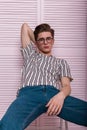 Modern sexy young man in fashionable youth wear in stylish glasses is resting on a chair near a wooden pink wall int the studio.