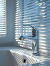 Modern silver faucet installed on white ceramic sink with sunlight creating striped shadows through window blinds in a clean Royalty Free Stock Photo