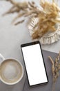 Modern tabletop with with smartphone mockup, cup of coffee, dried plants and notebook on textured white background. Flat lay,