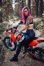 Modern tattooed hipster girl with bright pink hair sitting on her motocross bike in woods