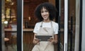 Millennial owner of modern cafe african american lady in apron holds digital tablet and standing near front door