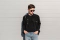 Modern trendy young man hipster in fashionable denim clothes in stylish sunglasses is resting near vintage building on street on Royalty Free Stock Photo
