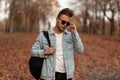 Modern trendy young man with a stylish hairstyle in black sunglasses in a denim jacket with a backpack posing in the autumn forest Royalty Free Stock Photo