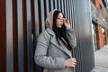 Modern urban stylish young woman model in fashionable long gray coat straightens stylish glasses near vintage metal wall in the Royalty Free Stock Photo