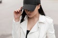 Modern urban young brunette woman in a fashionable black baseball cap in a stylish white leather jacket standing and looking down Royalty Free Stock Photo