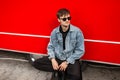 Modern urban young hipster man in fashionable denim clothes in trendy black sunglasses sits near a vintage red van on the street Royalty Free Stock Photo