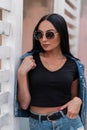 Modern urban young hipster woman in stylish black sunglasses stands and straightens a denim blue jacket near a vintage wooden wall Royalty Free Stock Photo