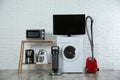 Modern vacuum cleaner and household appliances near white brick wall indoors