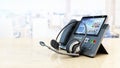 Modern VoIP phone and headset standing on wooden office table. Call center, marketing and technical support concept. 3D