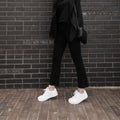 Modern woman in fashionable denim pants in leather stylish sneakers in trendy jacket with black handbag stands near brick wall in Royalty Free Stock Photo