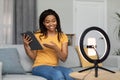 Modern work and hobby. Positive black female blogger shooting video blog, showing tablet, broadcasting in living room