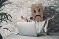 Modern worker with carton sad box on head open arms with desolation gesture in front of a laptop. Online crypto smart working Royalty Free Stock Photo