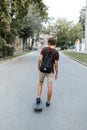 Modern young handsome man in summer shorts with a backpack in a hipster t-shirt is riding along the road on a skateboard Royalty Free Stock Photo