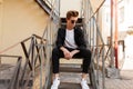 Modern young hipster man in trendy sunglasses in stylish black shirt in a t-shirt in elegant striped pants in sneakers sits