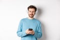 Modern young man in blue sweatshirt put on music on smartphone, listening songs in wireless headphones, white background Royalty Free Stock Photo