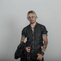 Modern young man hipster with tattoos in black trendy clothes with a jacket in round glasses posing in the studio near a vintage Royalty Free Stock Photo