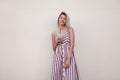 Modern young positive woman in a stylish long pink striped sundress stands near a vintage wall with a tasty milkshake in hands.