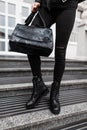 Modern young woman with a fashionable black leather bag in jeans in autumn stylish leather black boots is standing on the street.