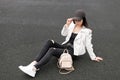 Modern young woman in white stylish leather jacket in a t-shirt in ripped jeans in white sneakers in a trendy  cap is resting Royalty Free Stock Photo