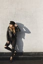 Modish blonde model girl in a leather jacket, ripped jeans, and black cap standing outdoors against the urban wall in