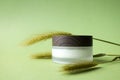 Moisturizer facial or eye cream in white frosted glass jar with wooden cup mockup and spikelet on green background