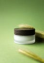 Moisturizer facial or eye cream in white frosted glass jar with wooden cup mockup and spikelet on yellow-green background